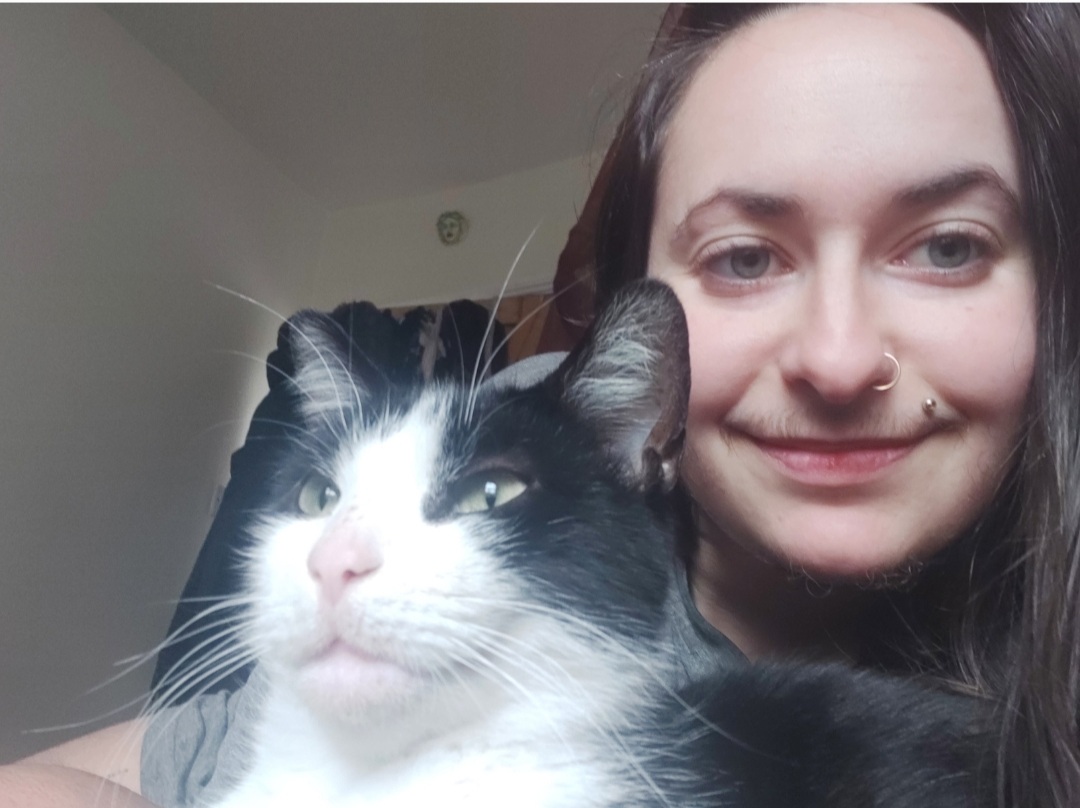 a photo of Sabina, a non-binary person withlong dark hair accompanied by their black and white cat
